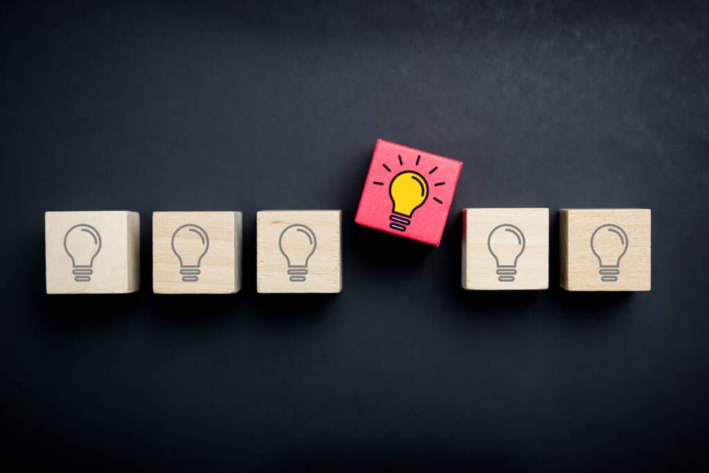 wooden blocks with light bulbs illustrated onto them lined up. One is out of the line, painted red, and the lightbulb is illuminated -- Learn proven branding differentiation strategies to make your small business stand out from the competition and become the preferred choice in a saturated market. -- Standing Out in a Crowd: Strategies for Differentiating Your Brand in a Saturated Market. -- your wp guy 