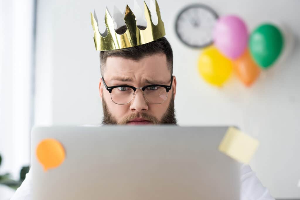 portrait of businessman with paper crown on head looking at laptop screen in office