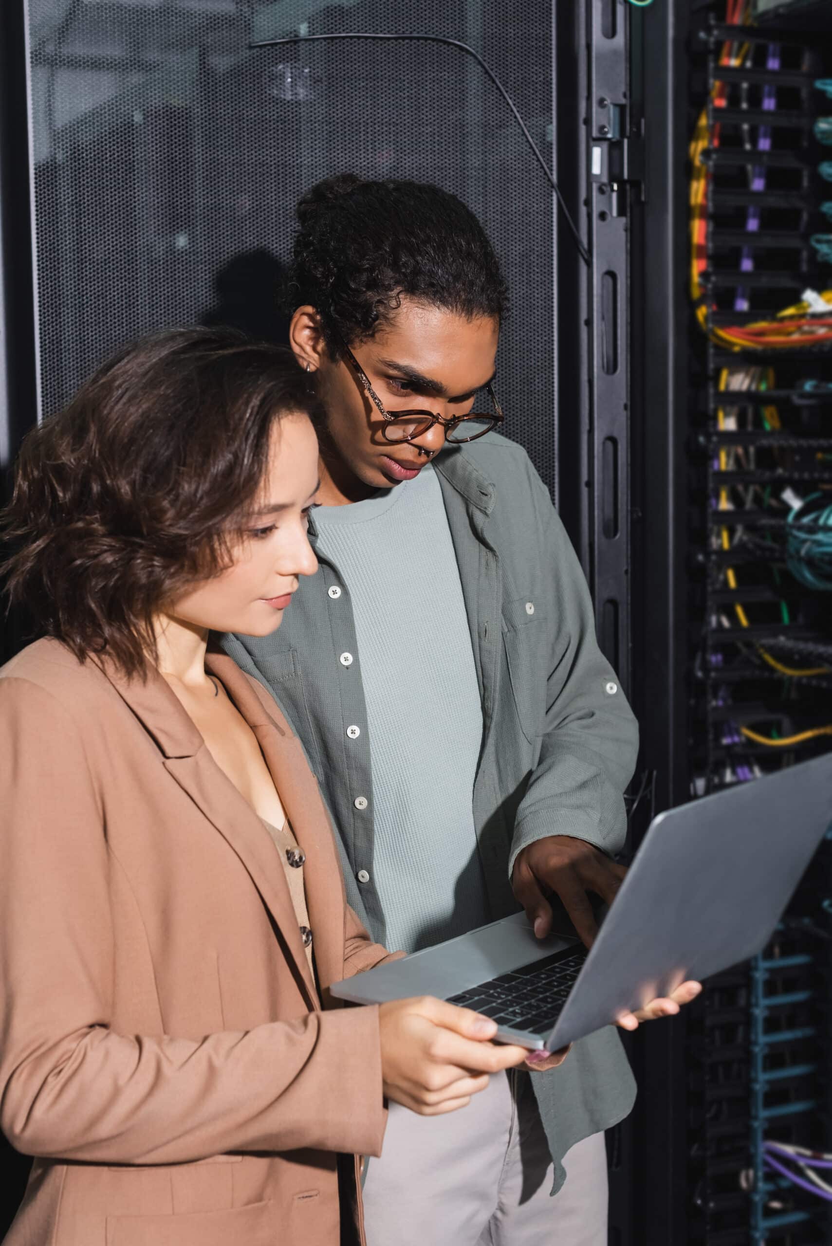 couple of multiethnic engineers looking at laptop while checking server in data center